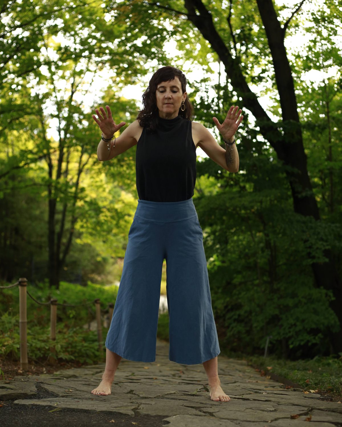 Julie Gazard practising qi gong barefoot on the same path, hands raised in front of her, under the trees.