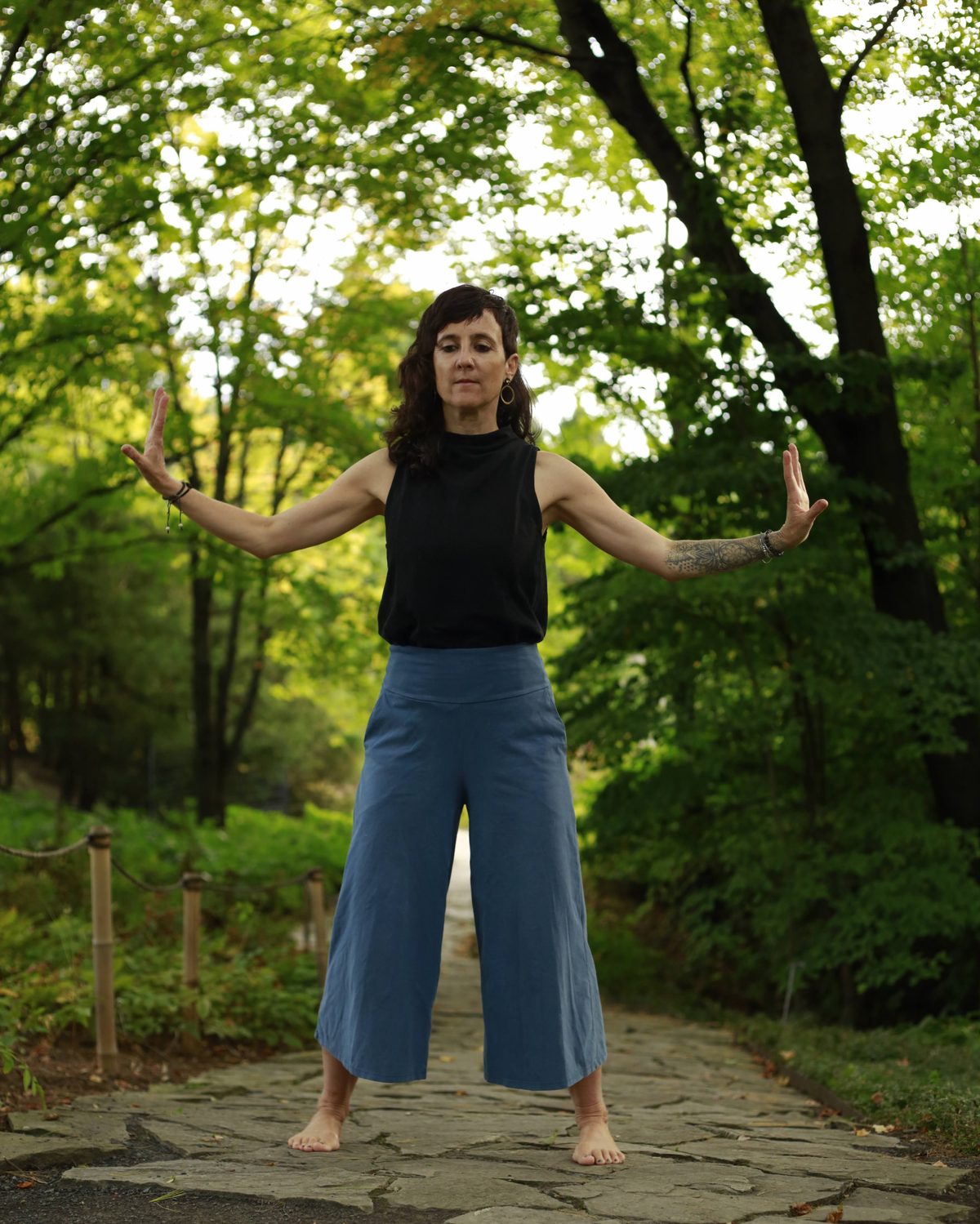Julie Gazard practising qi gong barefoot on a stone path in the Chinese Garden, arms open, under the trees.