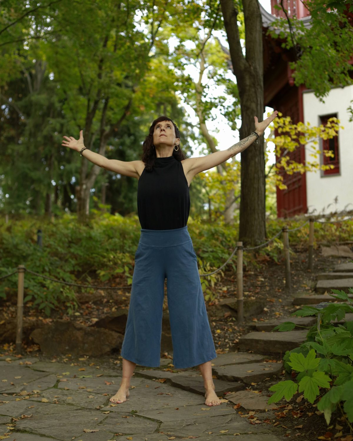 Julie Gazard practising qi gong barefoot on a stone path in the Chinese Garden, arms open, in front of a red and white pavilion.