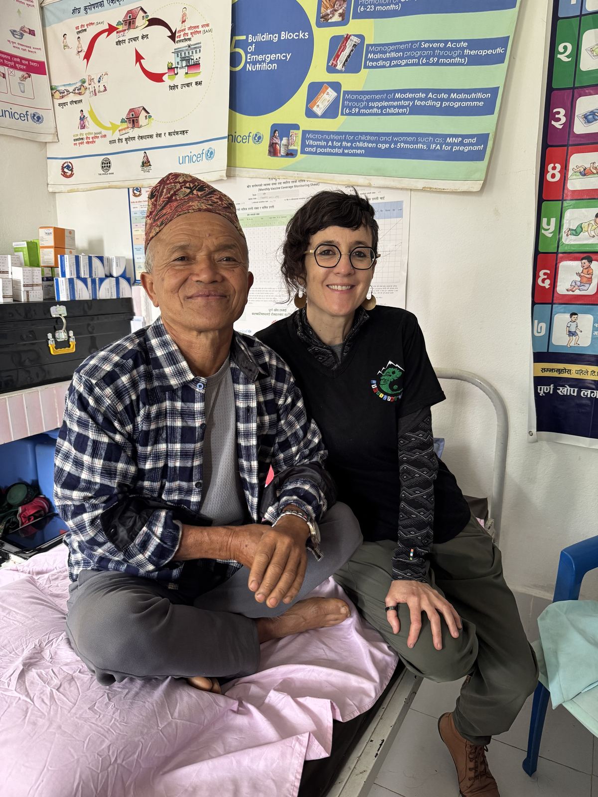 Julie Gazard, smiling, sitting on a bed next to a villager wearing a traditional hat, in Nepal.