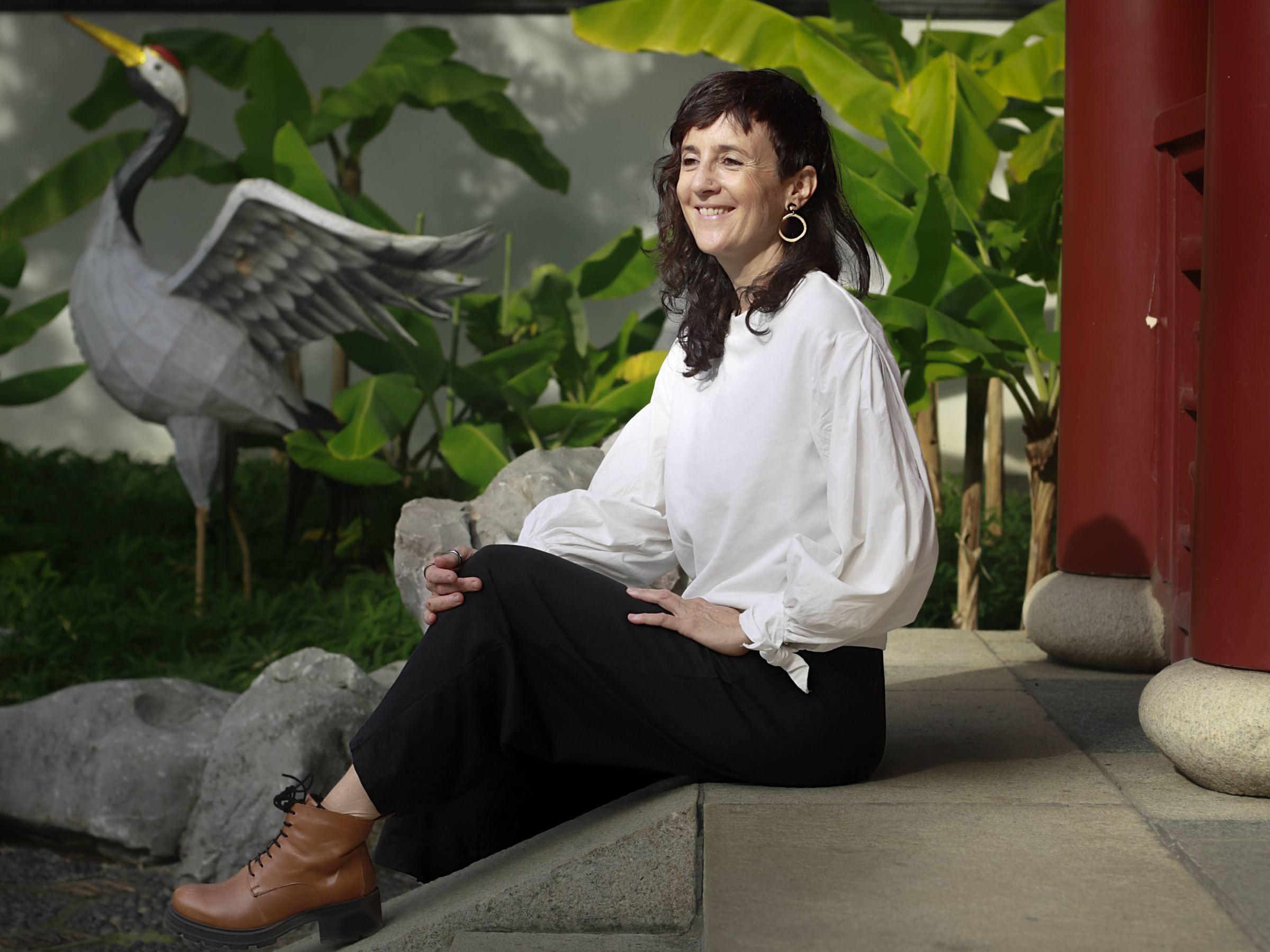Julie Gazard, en blouse blanche, assise au Jardin de Chine, près d'une grue de pierre.